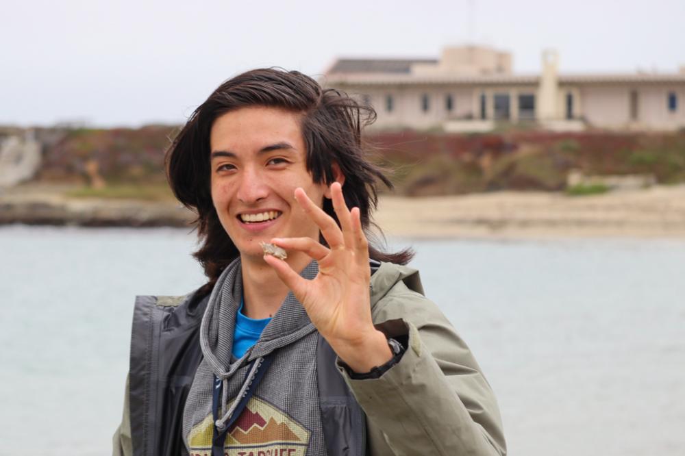 A student holding up a mole crab to the camera. In the background is Bodega Marine Laboratory