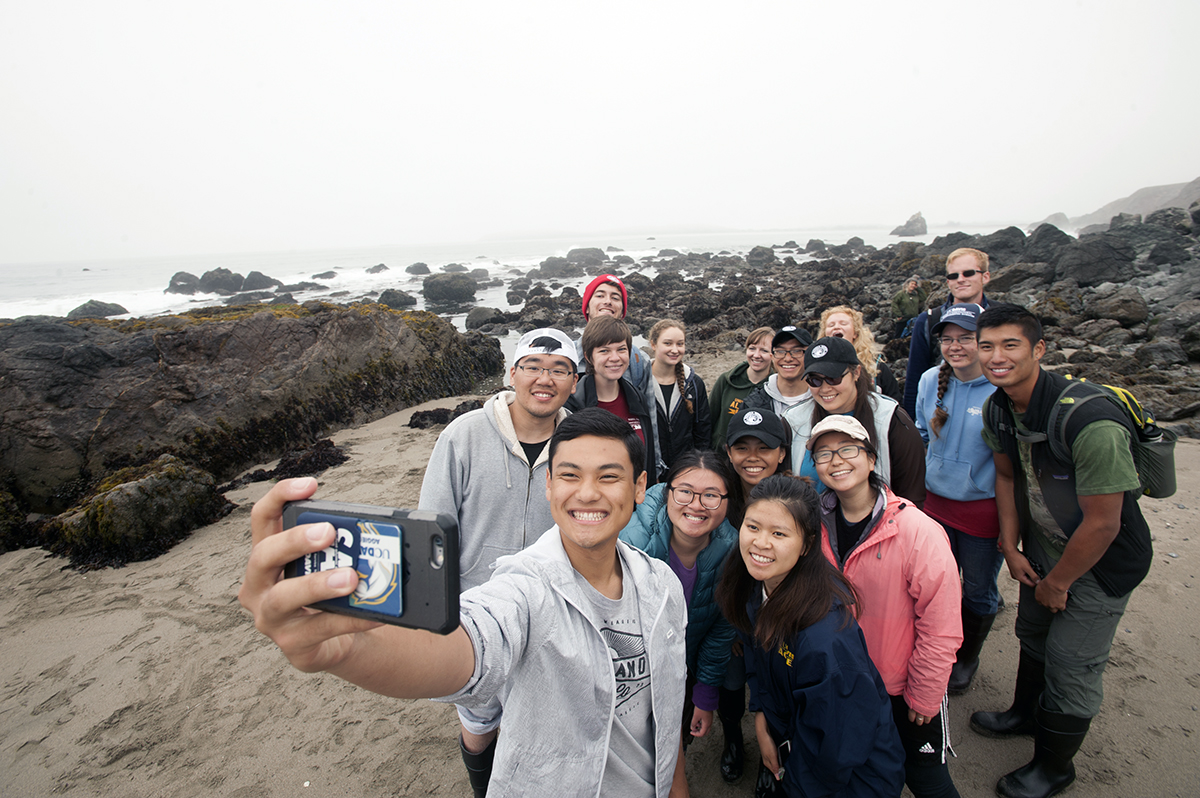 A group of undergraduate students gathered to take a group selfie, as the student front holds out a camera to take the photo. In the background is a rocky coastline and the ocean visible in the distance.