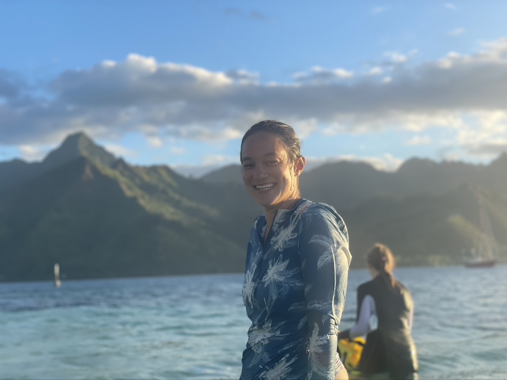 In the foreground, a person with long, dark hair that is pulled back and wearing a long sleeved shirt is standing waist deep in the ocean and smiling at the camera. In the background, another person stands in the water, facing away from the camera.