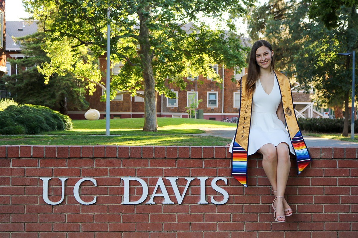 A student wearing a white dress and blue and gold UC Davis Graduate stole posed on a brick wall that says UC Davis on it.