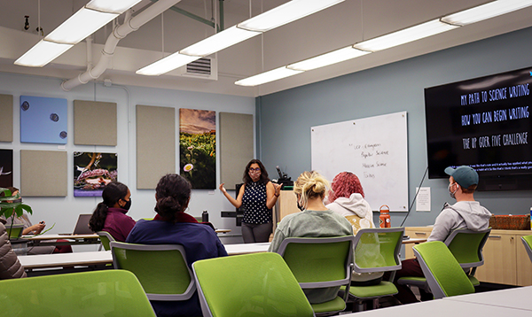 A person in a polka dot shirt and face mask standing in front of a group of students and gesturing while instructing a course on science writing.