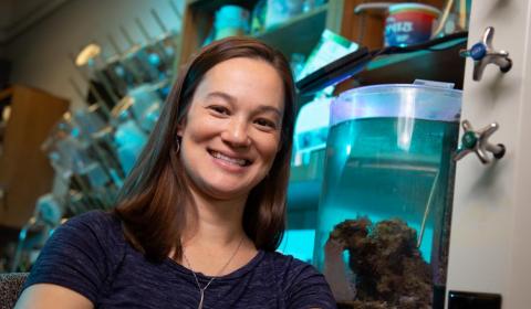A person with long brown hair smiling in front of lab equipment, including tanks full of blue liquid and marine organisms.