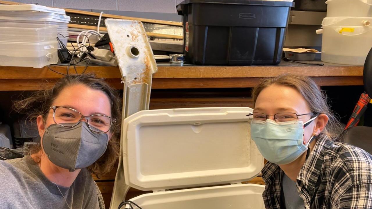 Two people posing in front of a cooler of research equipment smiling with masks on.