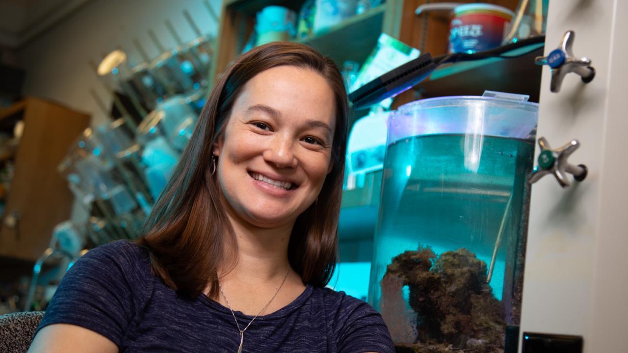 A person with long brown hair smiling in front of lab equipment, including tanks full of blue liquid and marine organisms.