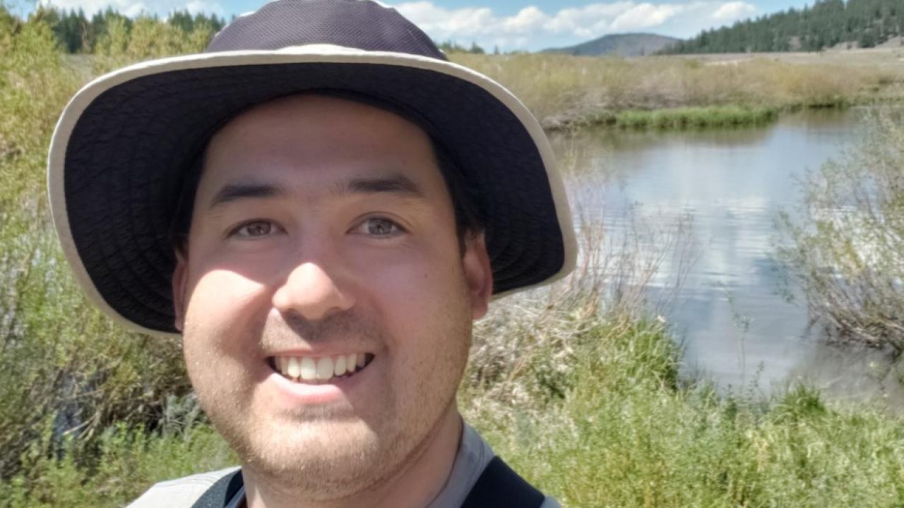 A smiling person wearing a sun hat and waders, with a marshy grass background and blue skies with fluffy white clouds.