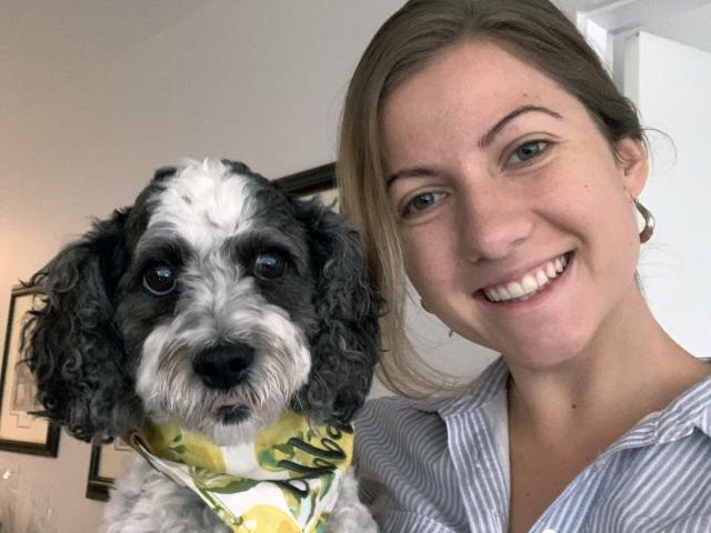 Photo of a person wearing a blue and white button down shirt with a black and white dog. The dog is wearing a yellow bandana around its neck.