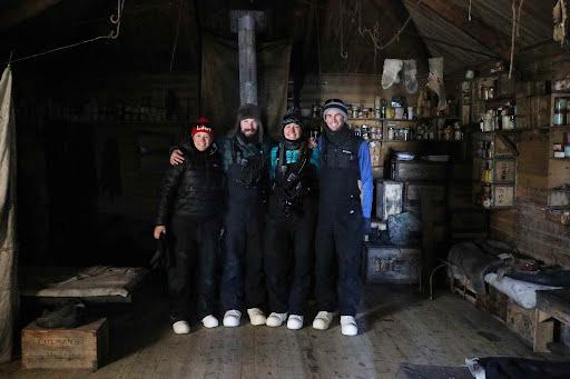 A group of four people standing with their arms around each other and smiling at the camera. They are all wearing black puffy overalls and hats with white shoes. Behind them is a wooden cabin interior with shelves full of kitchen equipment and food.