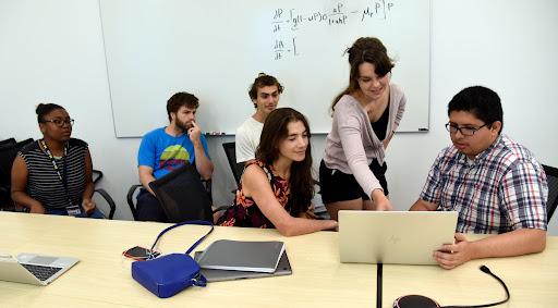A group of people gathered in a classroom setting in front of a laptop. One person leans in and points to something on the screen while the others watch.