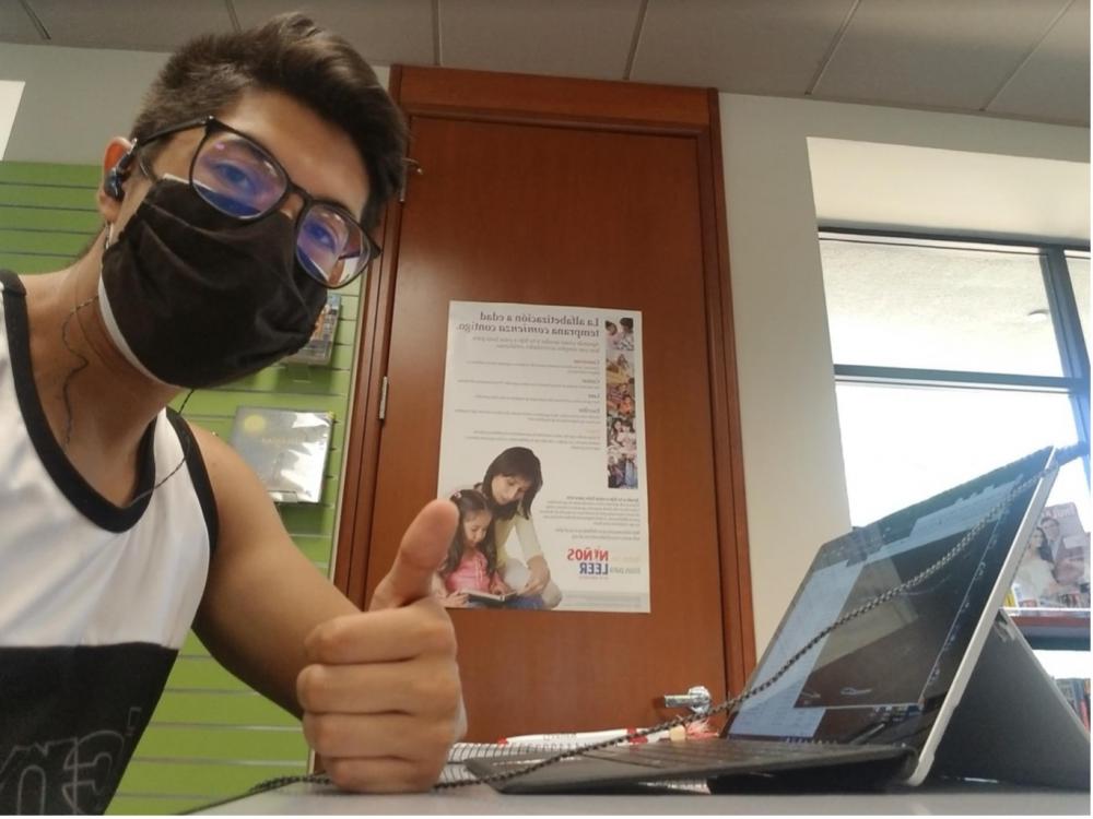 A person, wearing glasses and a mask, giving a thumbs up sign while sitting in front of a computer screen. In the background there is a door with a poster on it.