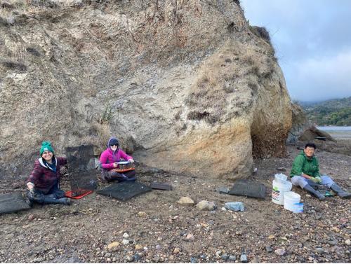 Three people sit on a rocky coastal shore, working with equipment as part of their research