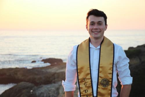 A man smiling in front of a sunset coastal background, wearing a UC Davis graduation stole