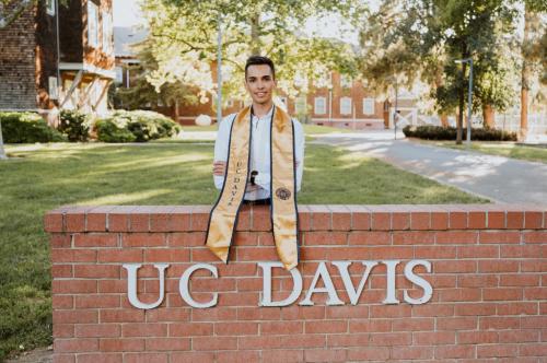 A person standing behind a brick sign that reads "UC Davis", wearing a UC Davis graduation stole