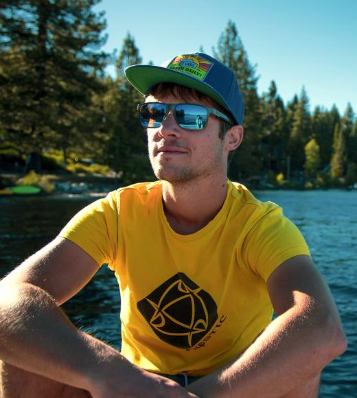 Florian Mauduit. Image shows a person dressed in a bright yellow shirt with a black graphic on it, wearing sunglasses and a dark colored baseball cap. Background is a body of water and evergreen trees.