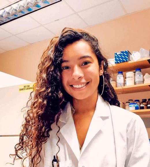 A person with long curly hair in a while lab coat, smiling at the camera