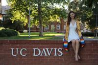 A woman sitting on a brick sign that says "UC Davis", wearing a white dress and a gold graduation stole