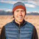 An image of Nicholas Trautman wearing a blue puffer vest and beanie, standing in front of a dry grassy landscape with blue skies and white clouds.